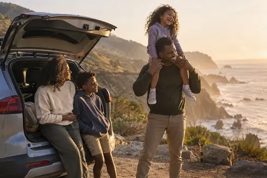 A family laughing at a coastal overlook beside their SUV at sunset