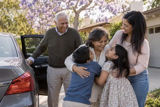 Grandparents hugging their grandchildren in a driveway beside their car